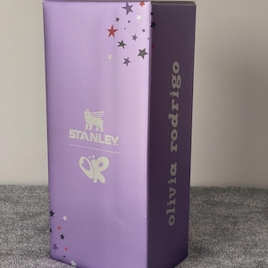 May include: A rectangular purple product box featuring the white Stanley logo with a winged bear icon and a white Olivia Rodrigo logo. The box is decorated with small red, white, and dark gray stars scattered across the top and bottom corners.