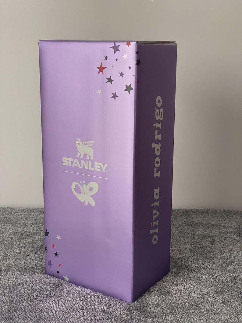 May include: A rectangular purple product box featuring the white Stanley logo with a winged bear icon and a white Olivia Rodrigo logo. The box is decorated with small red, white, and dark gray stars scattered across the top and bottom corners.