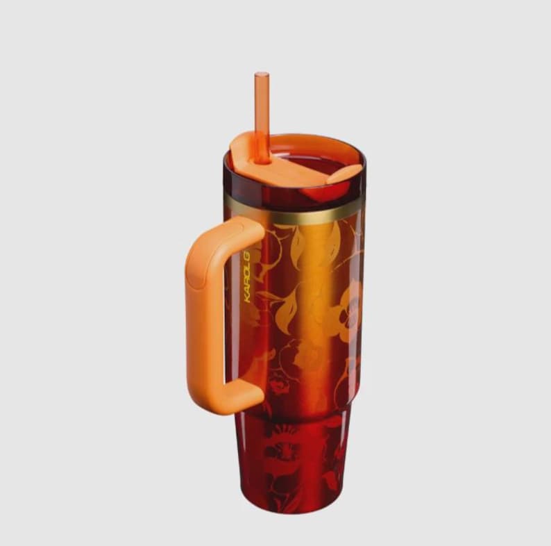 May include: A tall, translucent red tumbler with a gradient orange-to-red floral pattern, featuring a solid orange handle, a matching orange lid, and a vertical orange straw. The word KAROL G is printed vertically in small letters on the side.