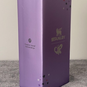 May include: A tall, rectangular purple cardboard box featuring the Stanley logo and a butterfly graphic. The box displays red, black, and white star patterns, along with the Instagram icon and the text @stanley_brand and @oliviarodrigo printed on it.