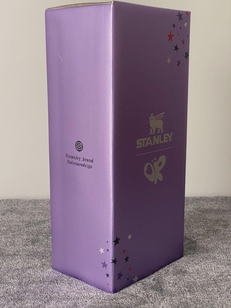 May include: A tall, rectangular purple cardboard box featuring the Stanley logo and a butterfly graphic. The box displays red, black, and white star patterns, along with the Instagram icon and the text @stanley_brand and @oliviarodrigo printed on it.
