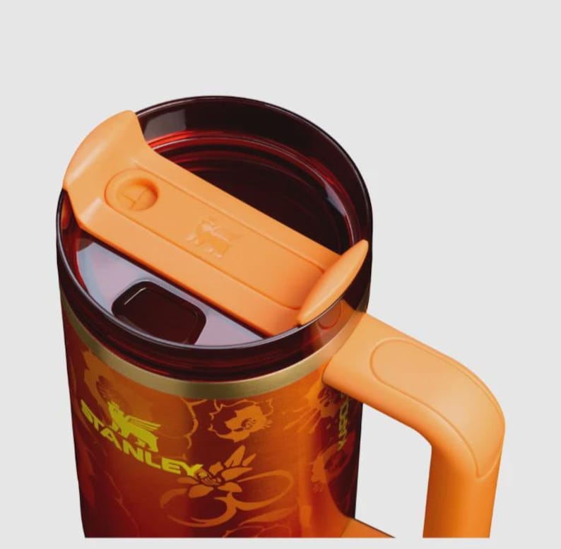 May include: A Stanley brand travel mug featuring a vibrant orange and red floral pattern on the exterior. The mug includes a translucent dark red lid with an orange sliding closure, a matching orange handle, and the Stanley logo printed in yellow.