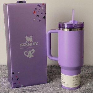 May include: A lavender Stanley tumbler with a matching handle, clear lid, and straw, standing next to its rectangular purple box. The box features the Stanley logo, a butterfly graphic, and scattered red, white, and purple star icons.