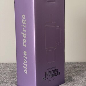 May include: A tall, rectangular purple product box featuring the text 'olivia rodrigo' vertically on the side. The front displays a line drawing of a tumbler with a straw and handle, labeled 'THE FLOWSTATE QUENCHER H2.0 TUMBLER' and '40 OZ | 1.18 L'.