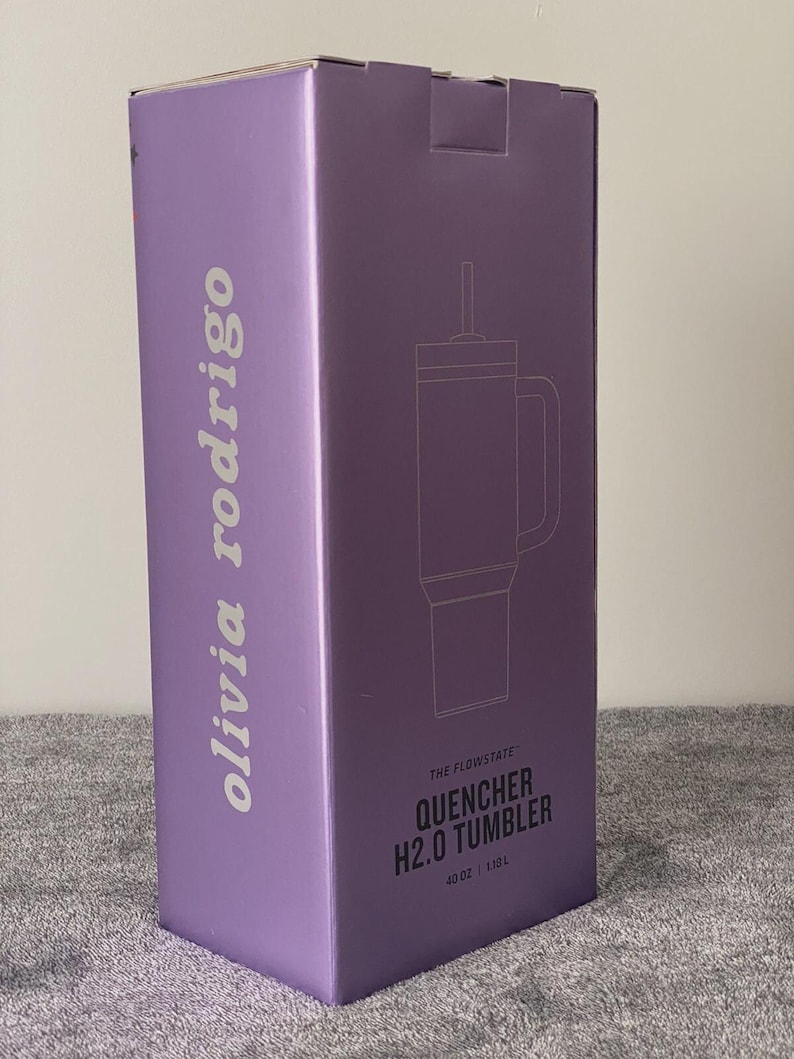 May include: A tall, rectangular purple product box featuring the text 'olivia rodrigo' vertically on the side. The front displays a line drawing of a tumbler with a straw and handle, labeled 'THE FLOWSTATE QUENCHER H2.0 TUMBLER' and '40 OZ | 1.18 L'.