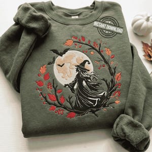 May include: An olive green crewneck sweatshirt featuring an embroidered design of a witch in a pointed hat standing before a large full moon. The scene is framed by dark tree branches, red berries, and orange leaves, with two small black bats flying.