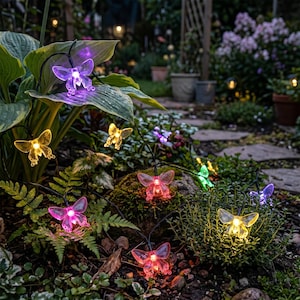 Könnte beinhalten: Eine dekorative Lichterkette mit schmetterlingsförmigen LED-Leuchten, deren durchscheinende Flügel in verschiedenen Farben wie Lila, Gelb, Pink und Grün leuchten. Die Schmetterlinge sind durch ein dünnes schwarzes Kabel verbunden und liegen im grünen Gartenlaub.