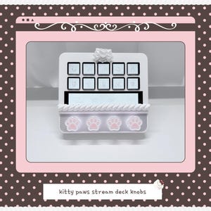 May include: A white stream deck console featuring ten square buttons with icons, a horizontal display screen, and a decorative base adorned with four light pink cat paw prints. A small white bow sits at the top center of the device frame.
