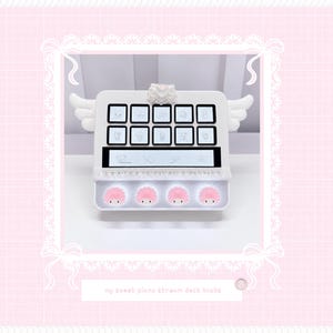 May include: A white stream deck featuring ten square buttons with character icons, a horizontal display screen, and four circular pink-haired character knobs. The device is adorned with white angel wings on the sides and a heart-shaped embellishment on top.