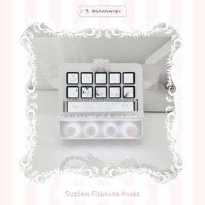 Puede incluir: Una vitrina blanca con diez pequeños pomos cuadrados, cada uno con un gráfico diferente en blanco y negro. La caja tiene un borde decorativo y el texto "Custom Fishcake Knobs" en la parte inferior. La parte superior de la caja está adornada con un lazo blanco.