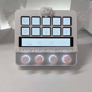 Peut inclure: Un contr&ocirc;leur rectangulaire blanc dot&eacute; de dix boutons carr&eacute;s avec des ic&ocirc;nes de personnages, un &eacute;cran d'affichage horizontal central et quatre boutons circulaires surmont&eacute;s d'accents en forme de rose rose. L'appareil est orn&eacute; d'ailes blanches.