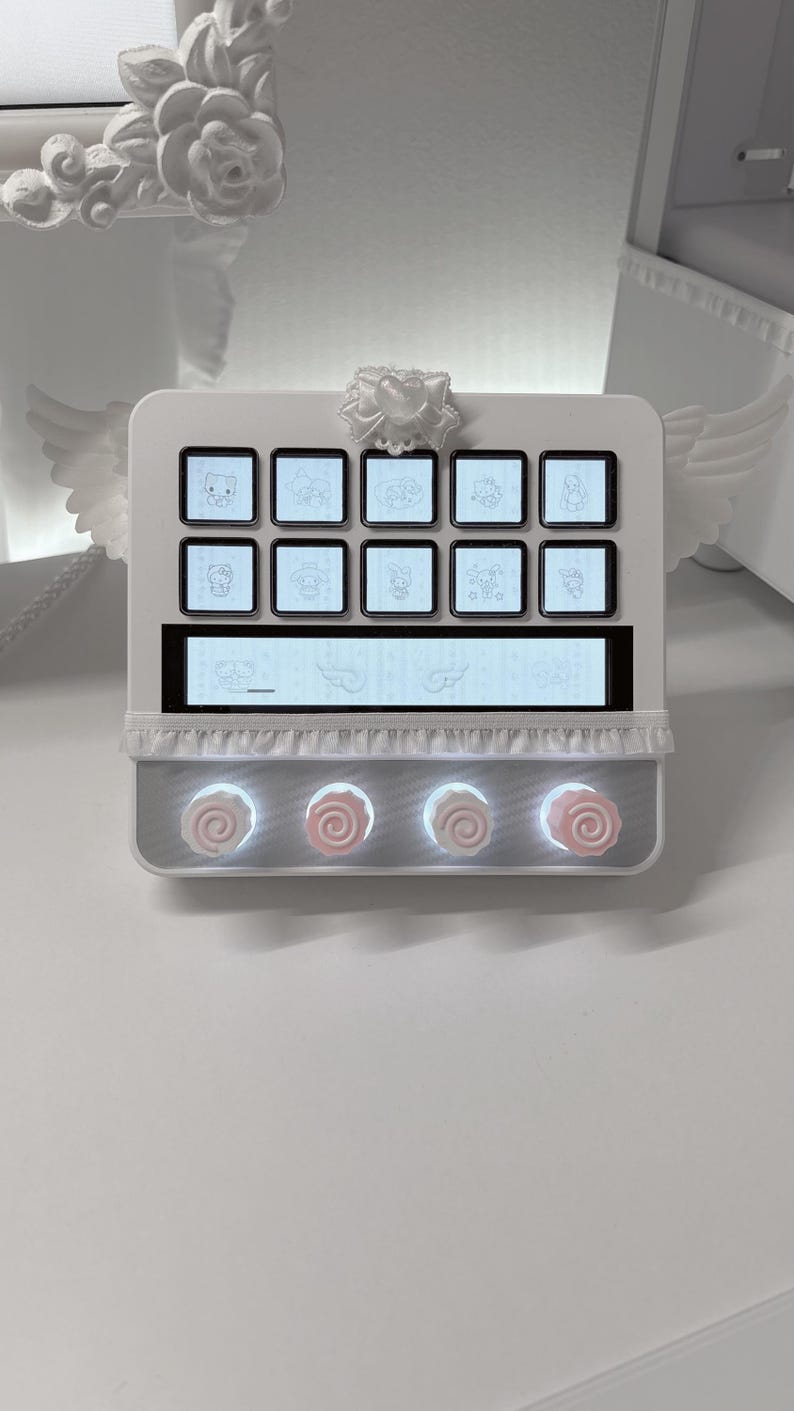 Peut inclure: Un contr&ocirc;leur rectangulaire blanc dot&eacute; de dix boutons carr&eacute;s avec des ic&ocirc;nes de personnages, un &eacute;cran d'affichage horizontal central et quatre boutons circulaires surmont&eacute;s d'accents en forme de rose rose. L'appareil est orn&eacute; d'ailes blanches.