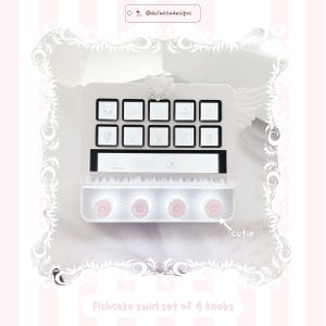 May include: A white rectangular device featuring ten square buttons with character illustrations, a horizontal display strip, and four circular knobs with pink fishcake swirl patterns labeled as cutie. The top includes a small white bow.