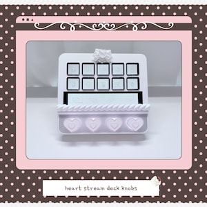 May include: A white stream deck console featuring ten square buttons with engraved icons, a central horizontal display screen, and four light pink heart-shaped knobs at the base. The device is topped with a small white bow-shaped decorative element.