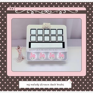 May include: A white stream deck featuring ten square buttons with light-colored icons, a horizontal display screen, and four circular knobs decorated with pink My Melody character faces. The device is accented with a white bow and lace trim.