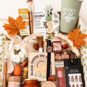 May include: A gift box containing a sage green travel mug with the text "BUT FIRST... Coffee", a coconut sheet mask, hand cream, essential oil, a journal with a rainbow graphic, fashion nails, tinted lip oil, body scrub, and decorative autumn leaves.