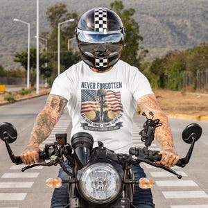 May include: A person wearing a white t-shirt with the words "Never Forgotten" and an American flag design, riding a black motorcycle. The person is wearing a black and white checkered helmet.
