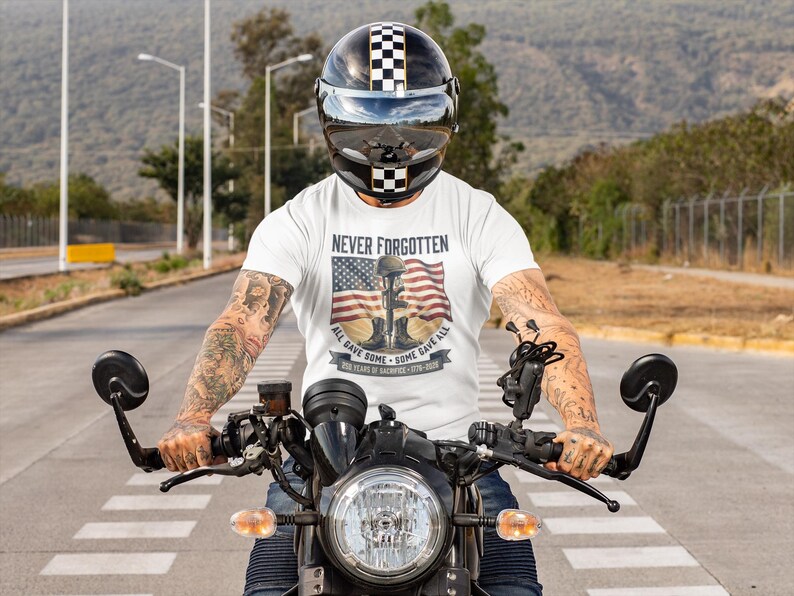May include: A person wearing a white t-shirt with the words "Never Forgotten" and an American flag design, riding a black motorcycle. The person is wearing a black and white checkered helmet.