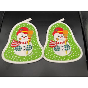 May include: Two bell-shaped oven mitts featuring a green background with white polka dots. Each mitt displays a snowman wearing a red hat and a red-and-white striped scarf, holding a broom. The word FRANCO is printed on the lower right.