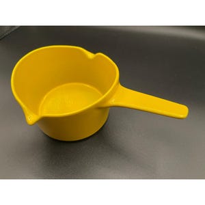 May include: A bright yellow sauce pan with a smooth, solid-colored finish. The vessel features a round, open-top design with a single integrated pouring spout on the side and a long, flat handle extending outward from the main body.