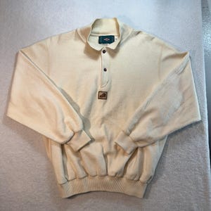 May include: A cream-colored long-sleeve pullover sweater featuring a mock neck collar with a two-button placket. The garment includes ribbed cuffs and a ribbed hem, with a small rectangular patch on the chest displaying a graphic of a shoe.