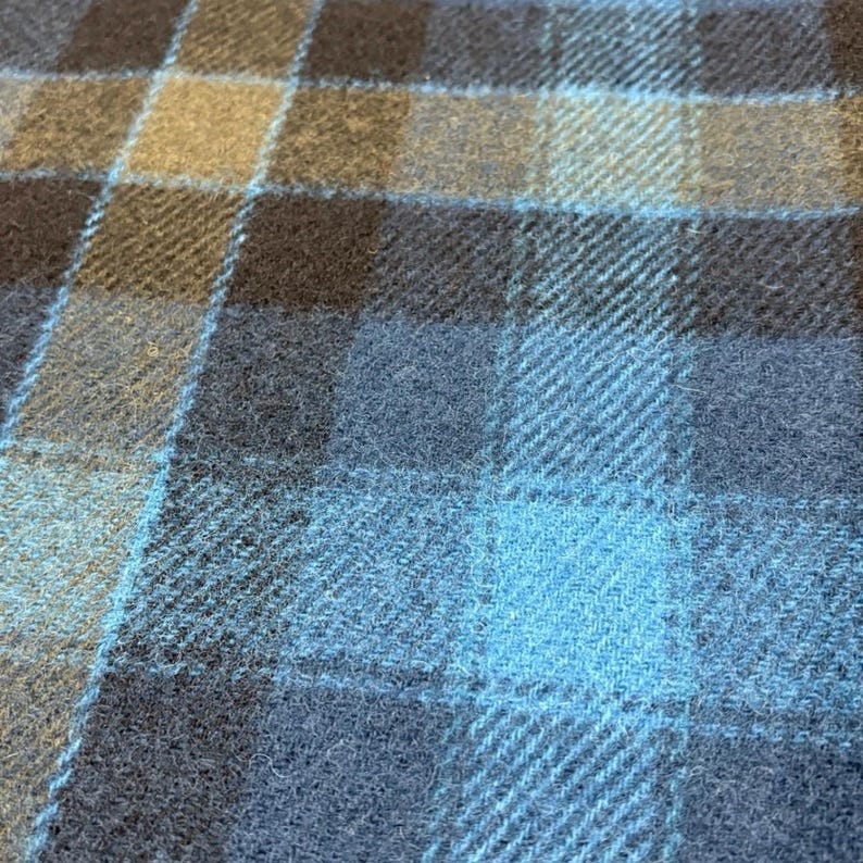 Pode incluir: Close-up de um tecido tecido com um padr&atilde;o xadrez. O design apresenta uma grade de linhas que se cruzam em tons de azul, marrom e bege. O tecido tem uma apar&ecirc;ncia texturizada, sugerindo l&atilde; ou material semelhante.