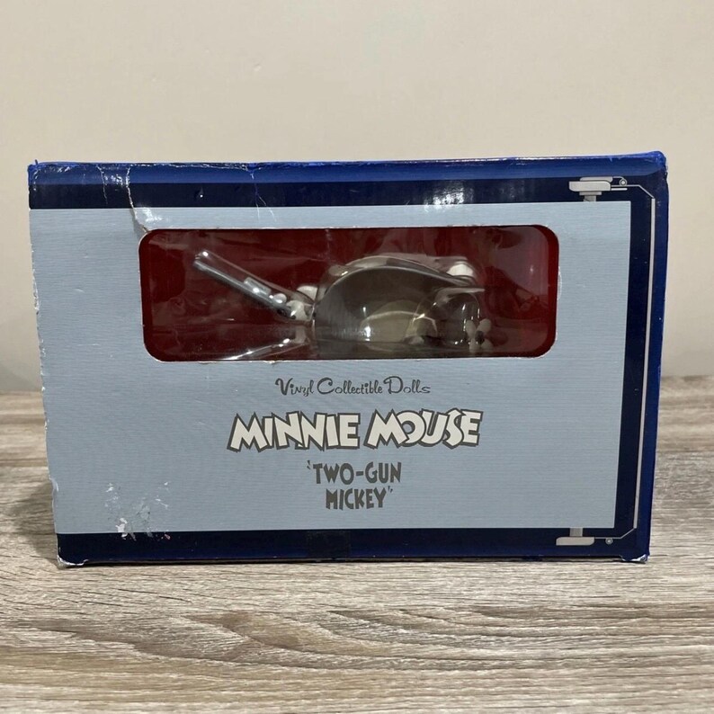 Pu&ograve; includere: Una bambola da collezione in vinile di Minnie Mouse in scatola. La scatola &egrave; blu e grigia con le parole "Vinyl Collectible Dolls" e "MINNIE MOUSE" stampate sul davanti. La bambola &egrave; etichettata "TWO-GUN MICKEY".