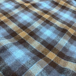 Pode incluir: Close-up de um tecido xadrez azul, castanho e bege. O padr&atilde;o &eacute; um design xadrez cl&aacute;ssico com linhas que se cruzam, criando quadrados e ret&acirc;ngulos. O tecido tem uma apar&ecirc;ncia macia e texturizada, adequada para roupas ou decora&ccedil;&atilde;o de casa.