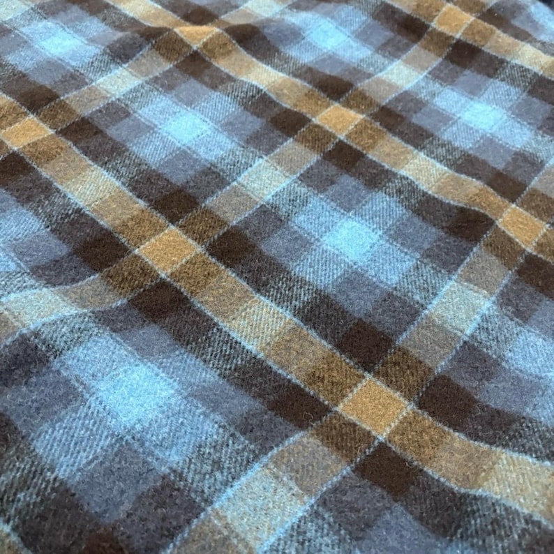 Pode incluir: Close-up de um tecido xadrez azul, castanho e bege. O padr&atilde;o &eacute; um design xadrez cl&aacute;ssico com linhas que se cruzam, criando quadrados e ret&acirc;ngulos. O tecido tem uma apar&ecirc;ncia macia e texturizada, adequada para roupas ou decora&ccedil;&atilde;o de casa.
