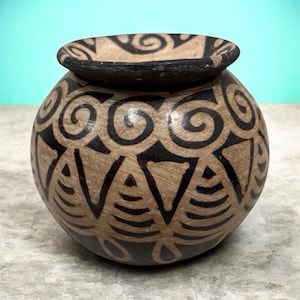 May include: A small, round, wooden bowl with a wide opening and a black rim. The bowl is decorated with black spiral and geometric patterns on a light brown background. The bowl has a unique, handcrafted appearance.