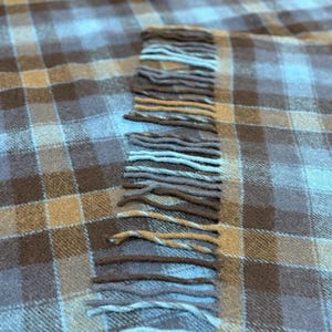 Pode incluir: Um close-up de um cobertor xadrez em tons de castanho, bege e azul. O cobertor tem uma borda com franjas com cores alternadas de castanho e azul. O cobertor &eacute; feito de um material macio.