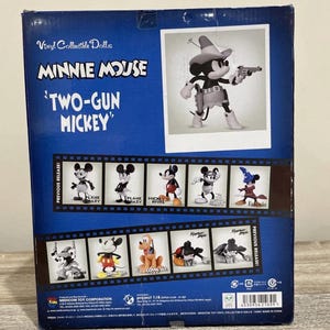 Pu&ograve; includere: Scatola blu per una bambola da collezione in vinile di Minnie Mouse, con "Two-Gun Mickey". La scatola include immagini di varie figure di Topolino e il testo "Vinyl Collectible Dolls".