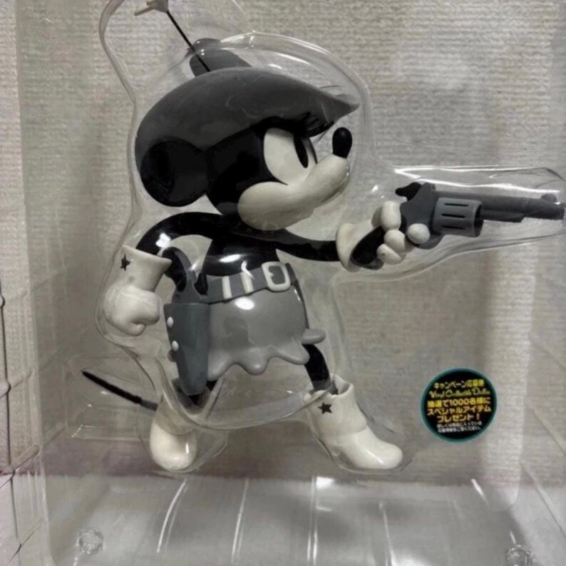 Pu&ograve; includere: Una statuetta nera, bianca e grigia di un topo dei cartoni animati in abito da cowgirl, con in mano un revolver grigio. Il topo indossa un cappello, una gonna e stivali. La statuetta &egrave; in una confezione di plastica trasparente.