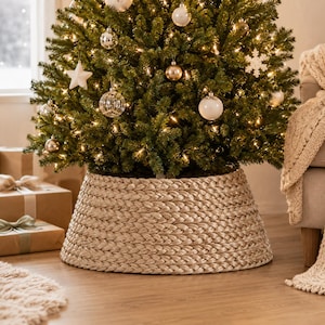 Puede incluir: Un cubrepies para árbol de Navidad trenzado en color champán con una silueta circular y cónica. La textura tejida presenta un brillo metálico y rodea la base de un árbol de Navidad decorado con adornos blancos, dorados y luces cálidas.