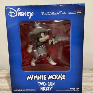 Pu&ograve; includere: Una bambola da collezione in vinile Disney di Minnie Mouse come "Two-Gun Mickey". La figura &egrave; nera, bianca e grigia, indossa un cappello da cowboy e tiene in mano una pistola. La scatola &egrave; blu con il logo Disney e informazioni sul prodotto.