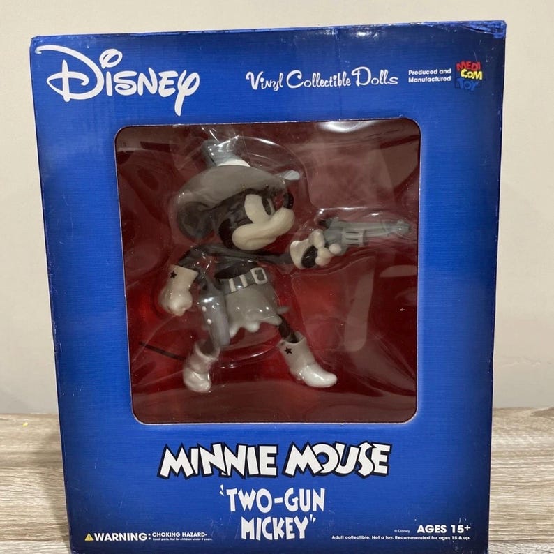 Pu&ograve; includere: Una bambola da collezione in vinile Disney di Minnie Mouse come "Two-Gun Mickey". La figura &egrave; nera, bianca e grigia, indossa un cappello da cowboy e tiene in mano una pistola. La scatola &egrave; blu con il logo Disney e informazioni sul prodotto.