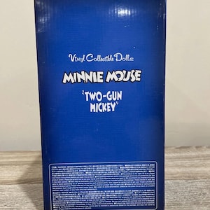 Pu&ograve; includere: Una scatola blu con la scritta "Vinyl Collectible Dolls", "MINNIE MOUSE 'TWO-GUN MICKEY'" e testo giapponese. La scatola &egrave; su una superficie di legno. &Egrave; un oggetto da collezione.