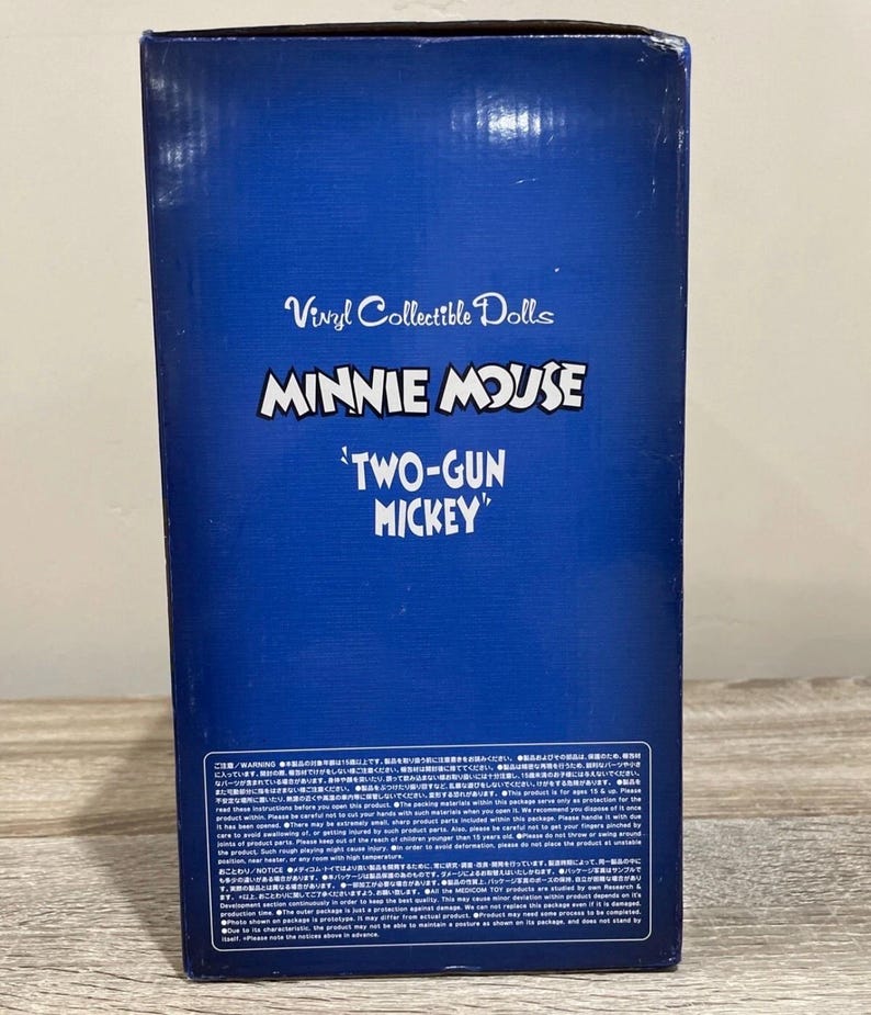 Pu&ograve; includere: Una scatola blu con la scritta "Vinyl Collectible Dolls", "MINNIE MOUSE 'TWO-GUN MICKEY'" e testo giapponese. La scatola &egrave; su una superficie di legno. &Egrave; un oggetto da collezione.