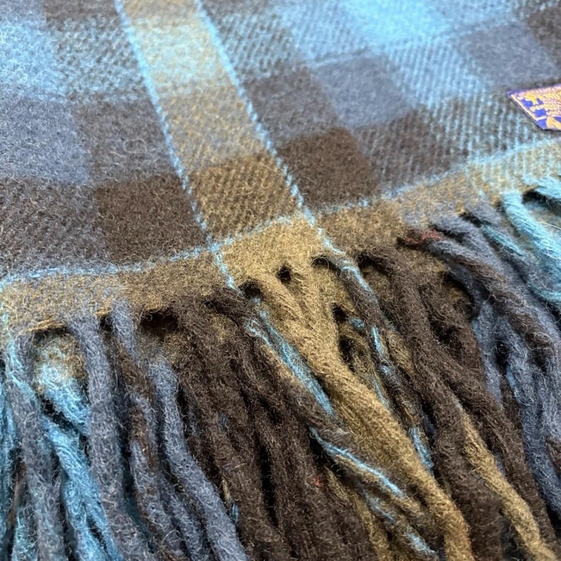 Pode incluir: Close-up de um cachecol xadrez azul, cinza e preto com franjas. O cachecol tem um padr&atilde;o xadrez cl&aacute;ssico em tons de azul, cinza e preto. A franja &eacute; um tom mais escuro de azul e preto.