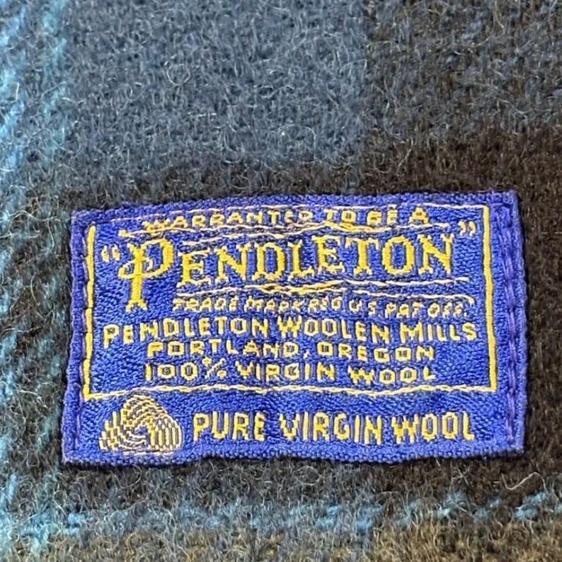 Pode incluir: Um close-up de uma etiqueta Pendleton azul e dourada. A etiqueta diz "Warranted to be a Pendleton" e inclui o texto "Trade Mark Reg. U.S. Pat. Off. Pendleton Woolen Mills, Portland, Oregon 100% Virgin Wool, Pure Virgin Wool."