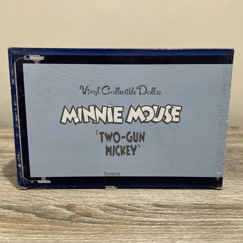 Pu&ograve; includere: Una scatola blu con le parole "Vinyl Collectible Dolls" e "MINNIE MOUSE 'TWO-GUN MICKEY'" stampate in bianco. La scatola ha un bordo blu scuro e si trova su una superficie marrone chiaro.