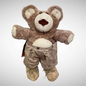 May include: A plush teddy bear with brown fur, a cream-colored muzzle, and dark brown nose. The bear wears beige shorts with metallic buttons and matching beige lace-up shoes. The bear has cream-colored fabric accents on the ears and paws.