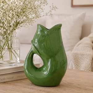 May include: A glossy, forest green ceramic vase shaped like a stylized fish with a curved tail forming a handle. The vase features a smooth, reflective surface and an open, wide-mouthed top, showcasing a distinct, sculptural silhouette.