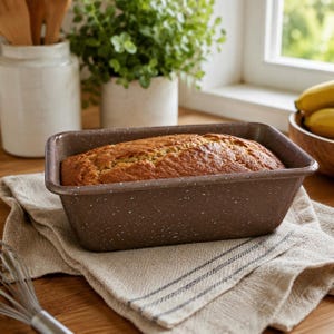 May include: A rectangular brown loaf pan with a white speckled finish containing a freshly baked golden-brown loaf of bread. The metal pan features a smooth rim and sits on a textured beige fabric cloth, showcasing a classic kitchen baking tool.