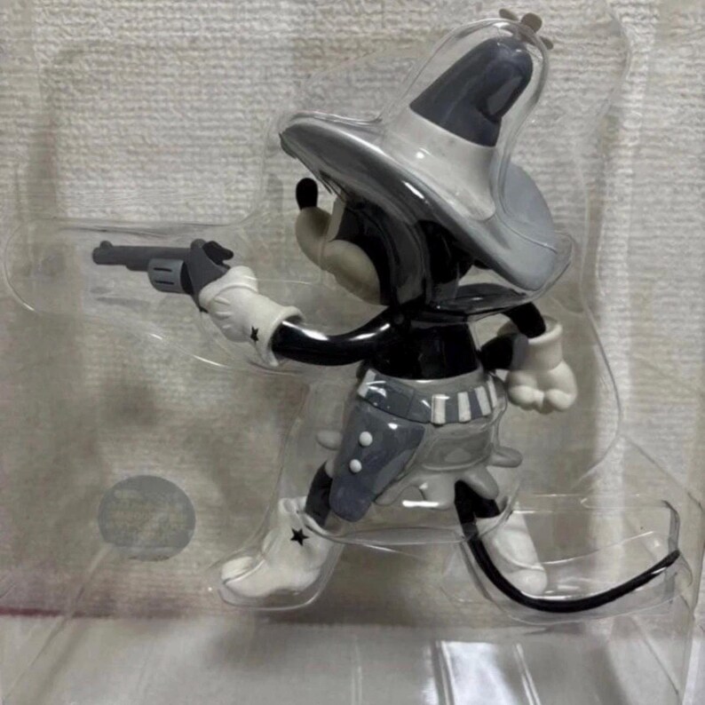 Pu&ograve; includere: Statuetta in bianco e nero di un topo dei cartoni animati in tenuta da cowboy, con una pistola. Il topo indossa un cappello da cowboy grigio, un gilet e stivali con una stella su ogni stivale. La statuetta &egrave; in una confezione di plastica trasparente.