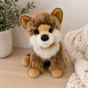 Peut inclure: Une peluche en forme de renard avec une fourrure douce marron et beige, dotée d'un poitrail et d'un museau blancs, d'oreilles pointues et d'un nez noir. L'animal en peluche est assis, avec une texture duveteuse et des yeux ronds et sombres.