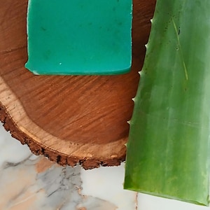 Puede incluir: Una pastilla de jabón cuadrada de color turquesa descansa sobre una rústica losa de madera circular. Una larga hoja verde de aloe vera con bordes espinosos está colocada a la derecha. El fondo presenta una superficie con vetas de mármol.
