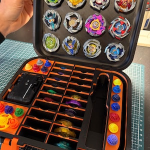 May include: A black and orange carrying case filled with colorful spinning tops. The case is open, revealing multiple compartments holding the tops and their accessories. The tops are various colors, including green, yellow, red, and blue.