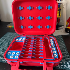 May include: Open red and blue plastic organizer with multiple compartments and slots. The interior features blue accents and various sized storage spaces. The organizer has a handle and latches for secure closure. The open case reveals a structured interior.