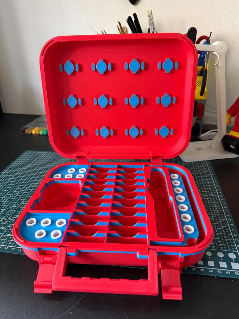 May include: Open red and blue plastic organizer with multiple compartments and slots. The interior features blue accents and various sized storage spaces. The organizer has a handle and latches for secure closure. The open case reveals a structured interior.